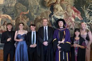 A group of LMH Tutors and students in formal clothes, celebrating while standing in front of a tapestry