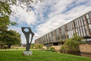 An abstract sculpture stands outside a 1960s modernist building with many windows, surrounded by green grass and trees