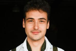 A portrait photo of a man with dark hair, wearing a white shirt and green braces