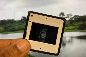 A close-up of a hand holding a photographic slide against a blurred backdrop of a calm lake and green trees. The slide contains an abstract image with geometric patterns. Promotional image for “Our Generous Mother,” an art exhibition by Onyeka Igwe.