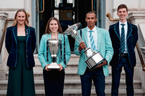 Two women and two men dressed in University of Oxford and University of Cambridge Blazers stand on some steps. One woman holds a large silver trophy