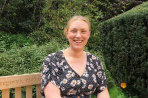 A woman with blonde tied-back hair wearing a navy dress is smiling while sitting on a bench surrounded by greenery