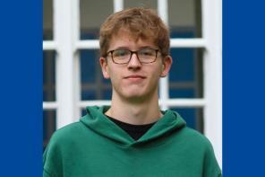 Jude Wood, who has short mid-brown hair and glasses. Jude is wearing a green hoodie and standing in front of a window