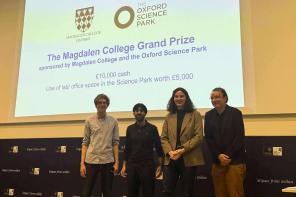 Four students stand on a stage in front of a screen saying: The Magdalen College Grand Prize