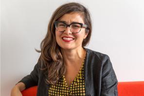 Professor Ana Domingos seated and smiling, wearing black-rimmed glasses, a black jacket, and a yellow and black polka-dot blouse, with a plain white wall in the background.