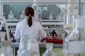 A photo of a woman wearing a white lab coat standing in a lab full of test tubes and other scientific equipment