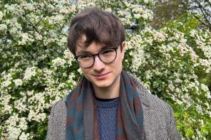 Leo Gitin, who has short dark brown hair and glasses, standing in front of a bush with lots of small white flowers
