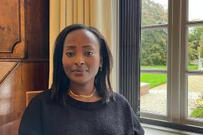 Dr. Makena Micheni sitting indoors by a large window with a garden view, wearing a black sweater and gold necklace, smiling softly at the camera