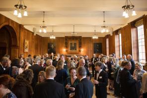 A large group of people standing and talking at an event in a wood-panelled room