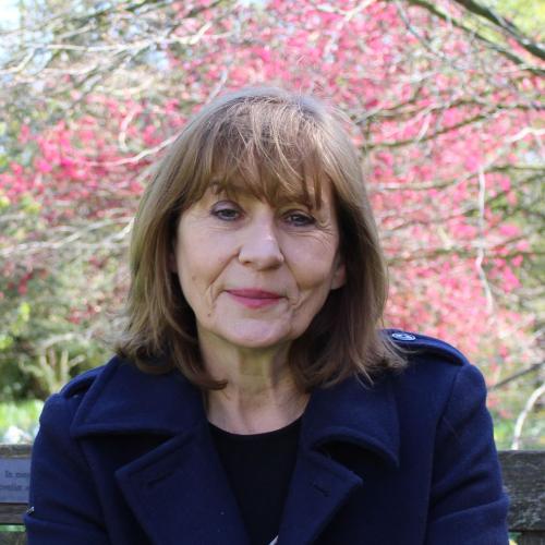 A woman with shoulder length dark brown hair wearing a dark blue coat sitting underneath a tree with pink blossom