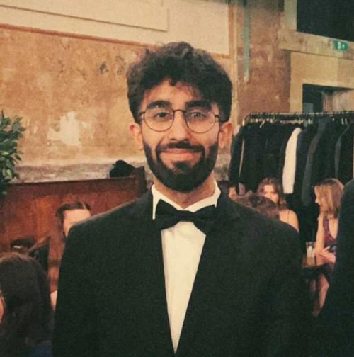 Photo of a young man with dark hair, a beard and glasses in a black suit