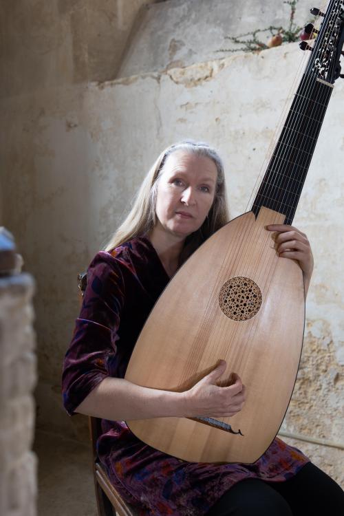 A woman with long silver hair sits holding a theorbo, similar to a lute