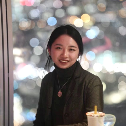 A woman with long tied-back dark hair sits at a table with city lights behind her