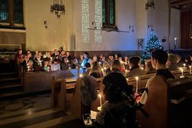 A candlelit carol service in the Lady Margaret Hall chapel. The choir is gathered in the front pews, holding lit candles and singing, while a speaker stands at the lectern. Congregants fill the pews, also holding candles. A lit Christmas tree glows to the right of the altar. The atmosphere is warm and reflective, with stained glass window reflections cast on the wall.