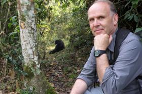 Professor Richard Wrangham sits to the right of the frame in a forest/jungle. In the background a gorilla sits in the middle of a path