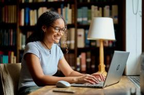 A young woman with tied-back curly brown hair sits at a desk in a library with her laptop
