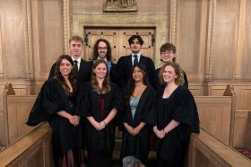 A group of students wearing black academic dress standing in front of some carved wooden doors in the LMH Chapel
