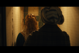A still from Onyeka Igwe's film 'A Radical Duet', showing two women wearing headscarves facing away from the camera and walking down a dimly lit corridor