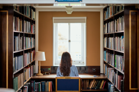 A student sits at a desk in a quiet library alcove, facing a window framed by bookshelves filled with books. Natural light streams in, creating a calm and focused study environment.