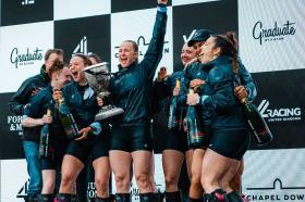 A group of 8 women and two men stand on a podium celebrating and holding a silver trophy after winning the Boat Race