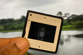 A close-up of a hand holding a photographic slide against a blurred backdrop of a calm lake and green trees. The slide contains an abstract image with geometric patterns. Promotional image for “Our Generous Mother,” an art exhibition by Onyeka Igwe.