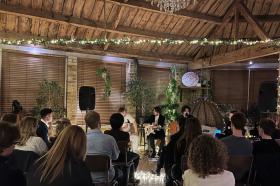 A group of young people sit in a wood-panelled room filled with fairy lights and greenery listening to a live band