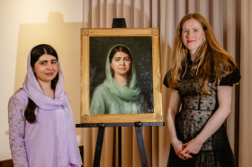 Malala Yousafzai, wearing a lavender purple outfit and headscarf, stands next to a portrait of herself alongside a woman in a black dress