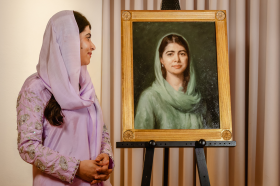 Malala Yousafzai, wearing a lavender purple outfit and headscarf, looks across at an oil portrait of herself
