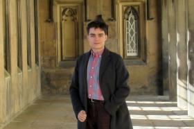 A young man in a long black coat standing in some stone cloisters