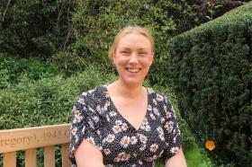 A woman with blonde tied-back hair wearing a navy dress is smiling while sitting on a bench surrounded by greenery