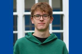 Jude Wood, who has short mid-brown hair and glasses. Jude is wearing a green hoodie and standing in front of a window