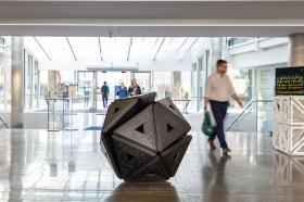 Schism (Black Oak) (2022) by Conrad Shawcross, displayed in the entrance hall of Oxford’s Mathematical Institute. The dark, geometric sculpture is formed from interlocking triangular wooden panels with inset metal bolts, creating a faceted, spherical form. People walk through the light-filled space around it, with signage for Shawcross’s Cascading Principles exhibition visible nearby.