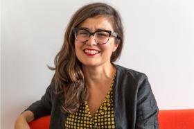 Professor Ana Domingos seated and smiling, wearing black-rimmed glasses, a black jacket, and a yellow and black polka-dot blouse, with a plain white wall in the background.