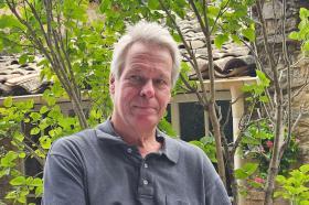 Professor Jochen Koenigsmann sitting outdoors in front of a stone building and leafy green plants, wearing a dark grey collared shirt