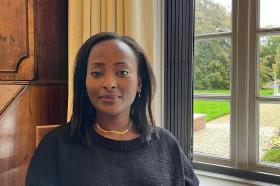 Dr. Makena Micheni sitting indoors by a large window with a garden view, wearing a black sweater and gold necklace, smiling softly at the camera