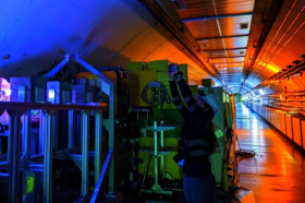 A researcher wearing safety gear works inside CERN’s HiRadMat irradiation facility, standing beside large industrial equipment under vibrant blue and orange lighting in a long, tunnel-like experimental hall