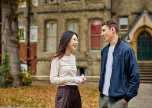 Two students, one male and one female, stand talking animatedly outside of a victorian-style brick building