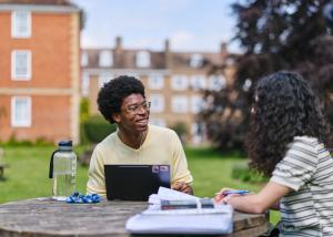 Two students sitting at a wooden table in the LMH gardens with books and laptops in front of them