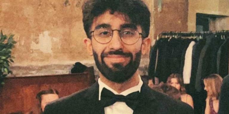 Photo of a young man with dark hair, a beard and glasses in a black suit