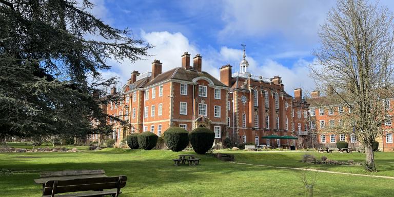 A red brick building set in gardens featuring green grass and large trees, with a blue sky