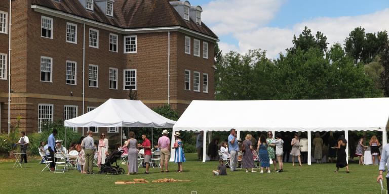 Photo of people gathered around a large white marquee on a green lawn at a garden party