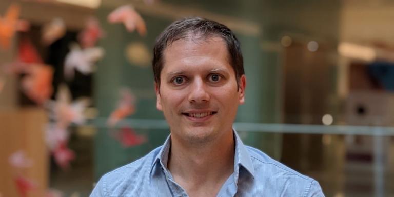 Oliver Pambos, who is wearing a pale blue shirt and standing in an atrium