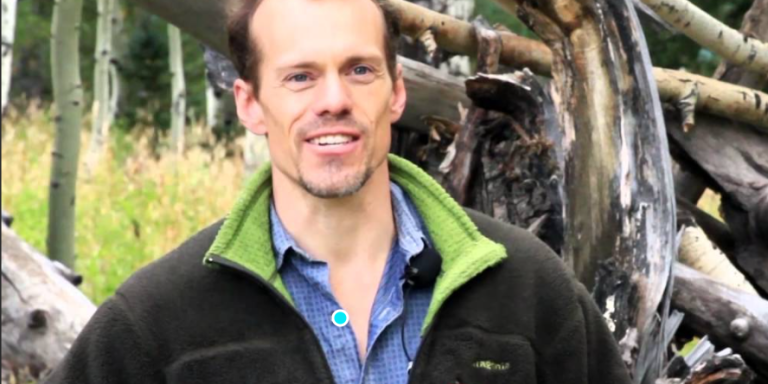 A man with short brown hair, a blue shirt and dark fleece stands in a forest