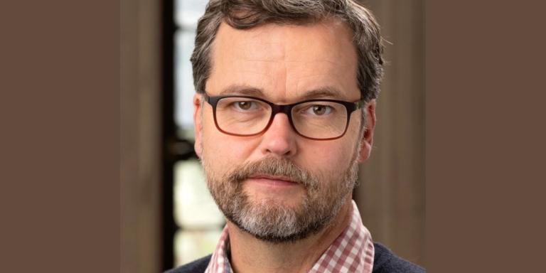 A man with short brown hair and a beard with rectangular-framed glasses looks at the camera