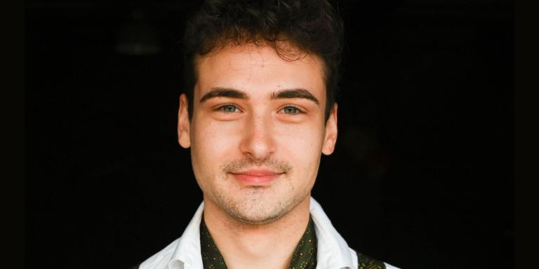 A portrait photo of a man with dark hair, wearing a white shirt and green braces