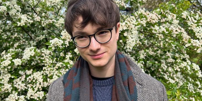 Leo Gitin, who has short dark brown hair and glasses, standing in front of a bush with lots of small white flowers