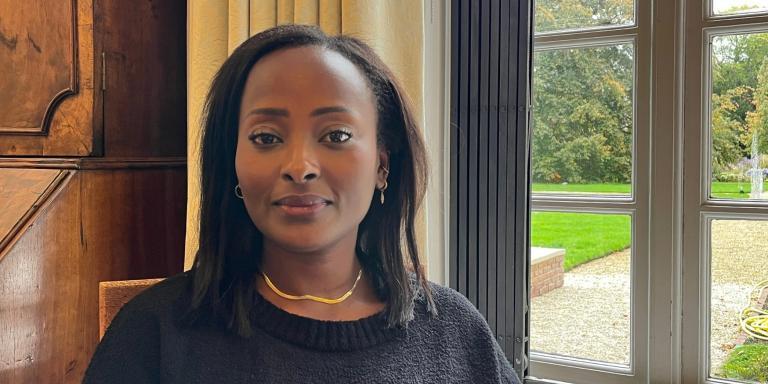 Dr. Makena Micheni sitting indoors by a large window with a garden view, wearing a black sweater and gold necklace, smiling softly at the camera