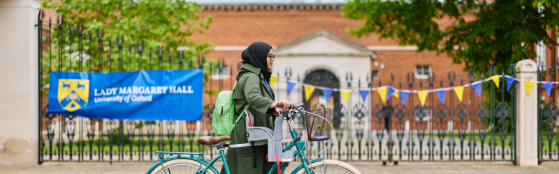 A lady walking a bicycle with a child seat past the front gates of Lady Margaret Hall, University of Oxford, with the College sign, wrought-iron gates, and historic red-brick buildings in the background