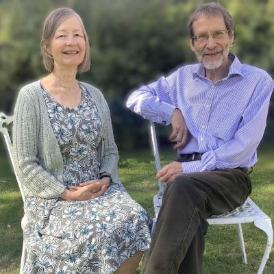 Sarah Gould, wearing a green floral dress and muted green cardigan and Andy Watson, who is wearing a pale purple shirt, sit on white garden chairs on a green lawn 