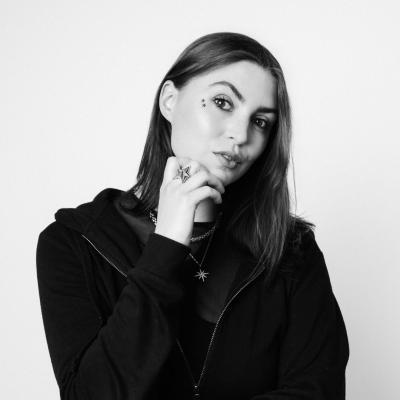 Black and white photo of Emmeline Armitage, a woman in her mid-twenties with long dark brown hair. She is looking at the cameral with her eyebrow raised.